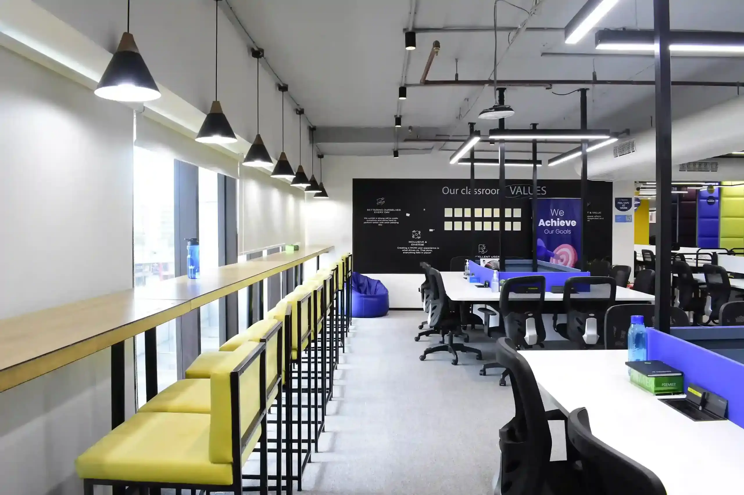 Modern coworking space with a window-facing wooden bar table, yellow bar stools, and rows of office desks under pendant lights.