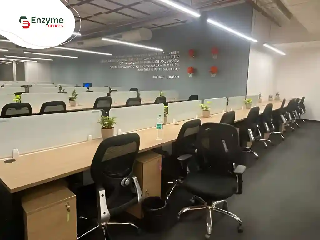 Open office workspace with long desks, chairs, partitions, plants, and overhead lighting
