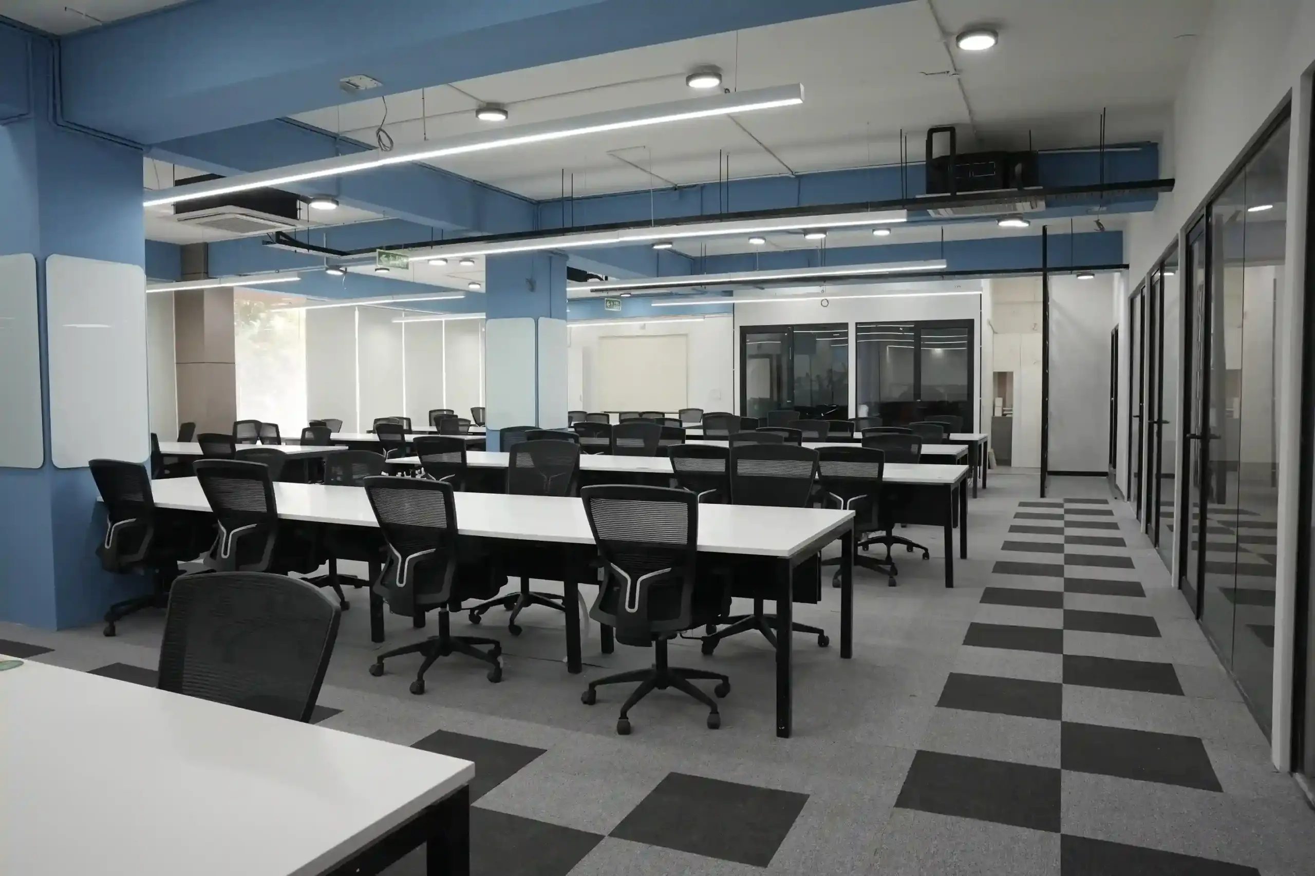 Open-plan office with rows of white desks, black chairs, blue ceiling beams, and a distinctive checkered carpet runner.